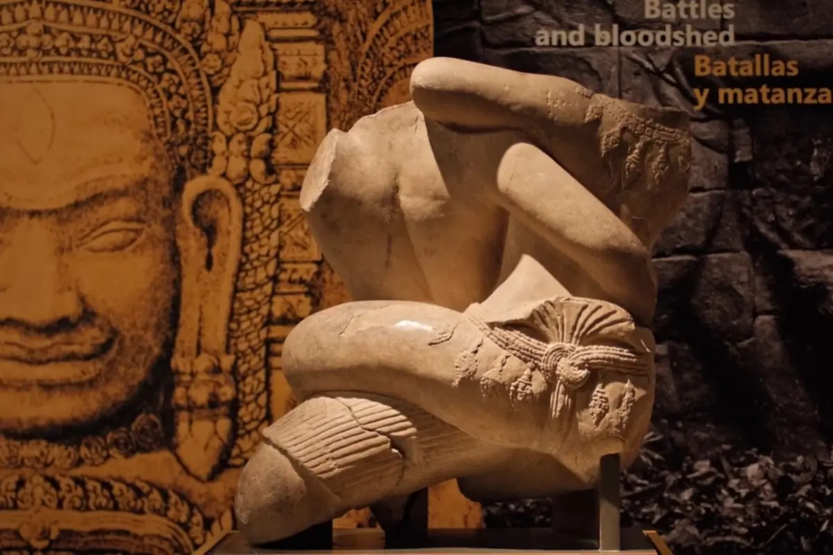En stenskulptur af to khmer-brydere, den ene bøjet over den anden, udstillet foran et stort udskåret ansigt på en Angkor-udstilling