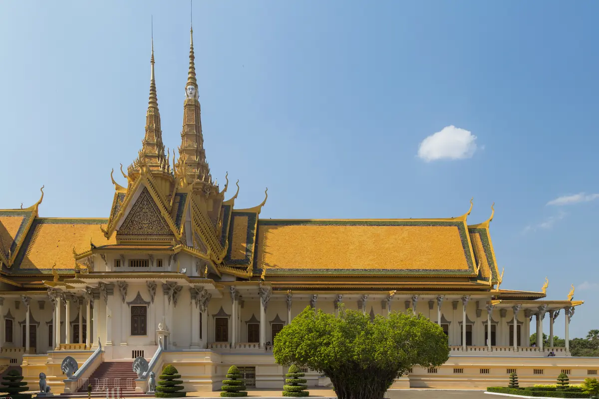 Trůnní síň Královského paláce v Phnom Penhu se zlatou střechou, se dvěma vysokými věžemi, bílými sloupy a před ní banánovníkovcem pod modrou oblohou